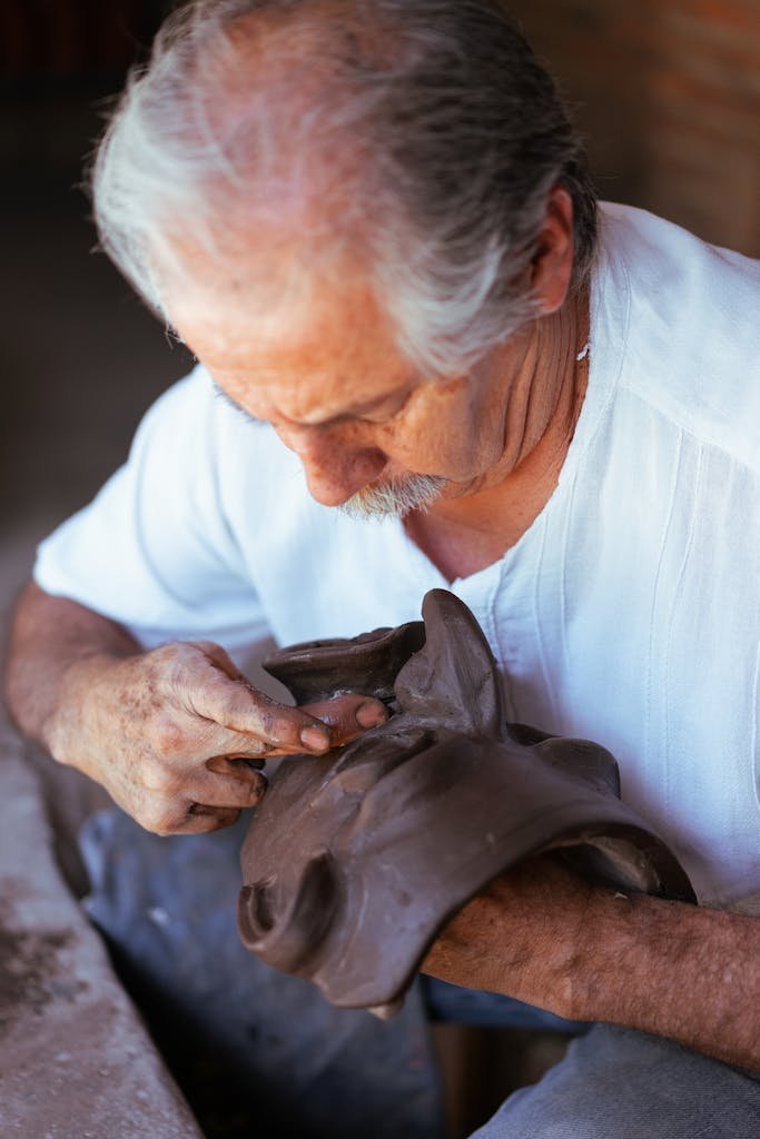 Elderly man shaping clay mask with precision in a traditional workshop setting.